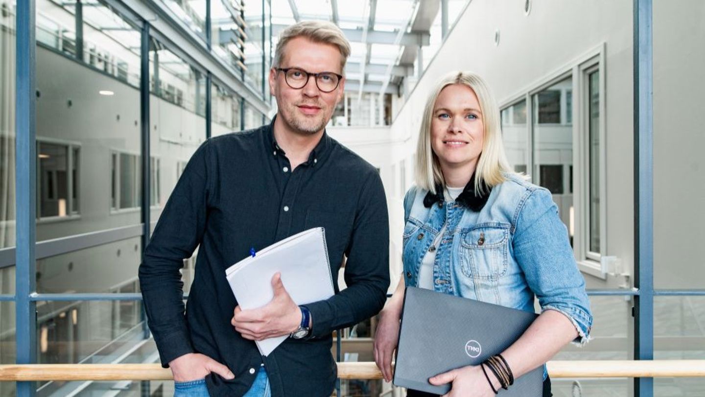 Bildet viser to kolleger som står ved siden av hverandre. De har blokk og PC i hendene og ser rett i kameraet. 