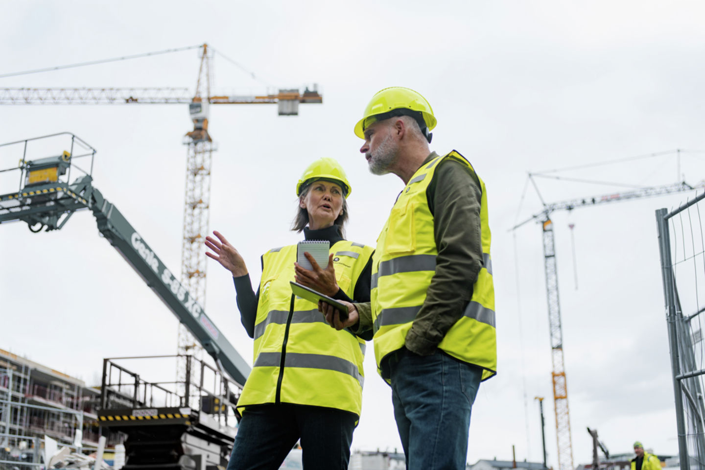 Mann og kvinne med hjelm og refleksvest står og snakker på en byggeplass med to heisekraner i bakgrunnen