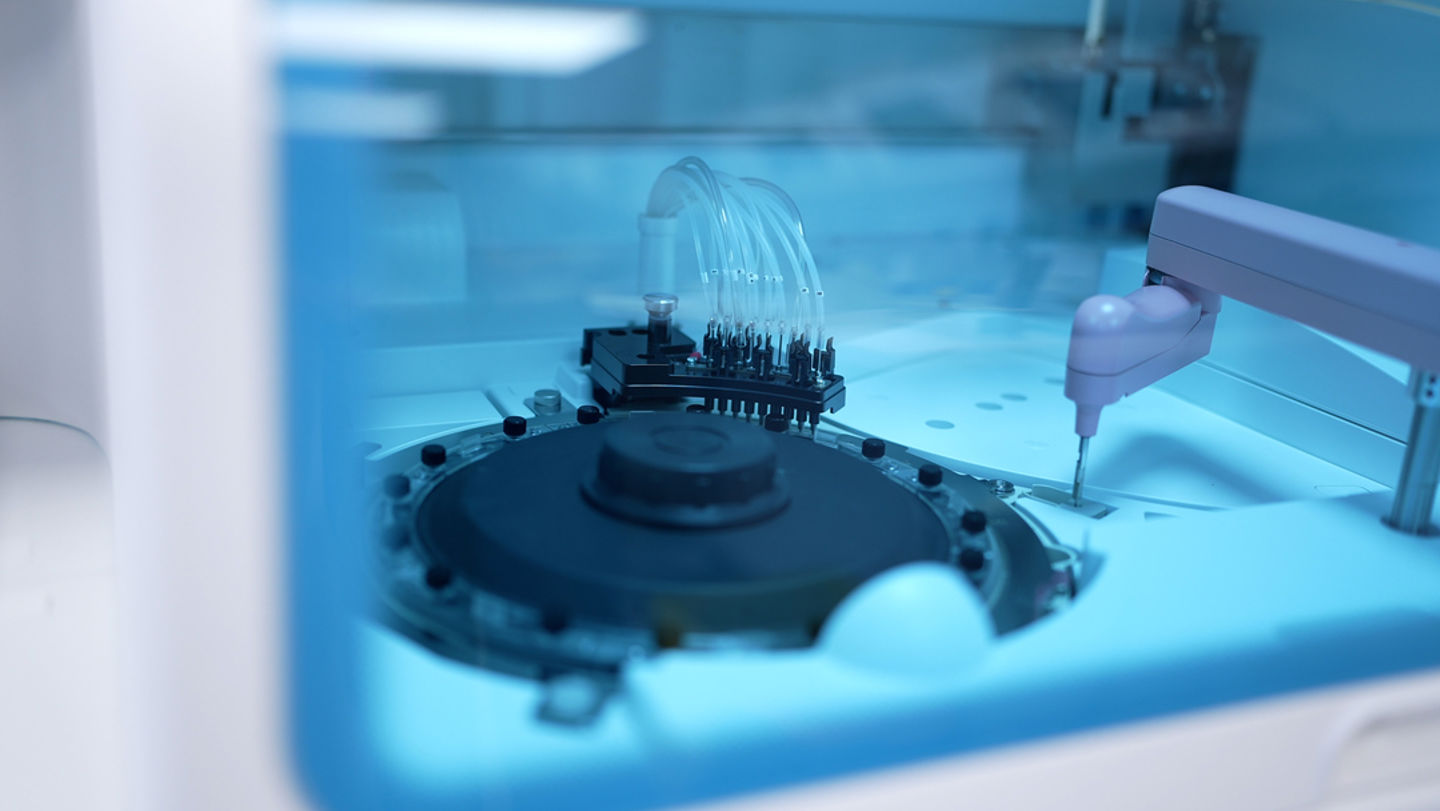 Nærbilde av en automatisert laboratorie- eller industrimaskin med robotarm og prøvehåndteringsutstyr.
