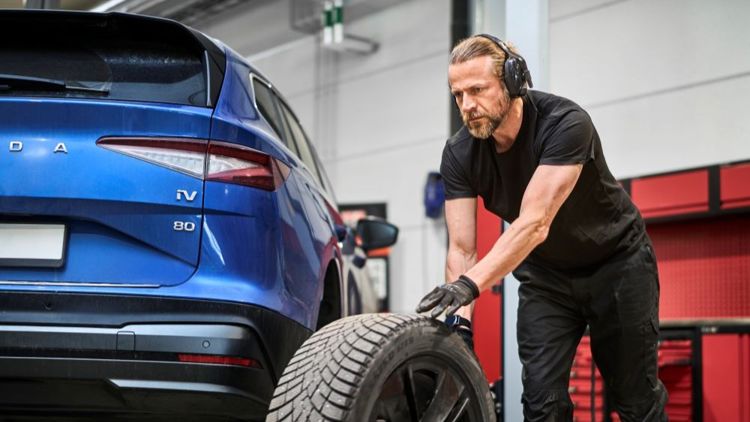 Bildet viser en mann som jobber på et bilverksted. Han triller et hjul bort til en blå bil. 