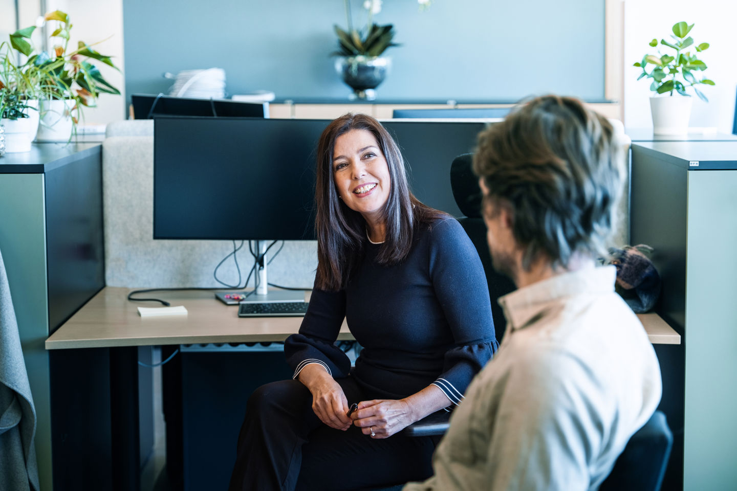 To personer sitter mot hverandre i et kontorlandskap. Vi ser bakhodet til en mann som ser på en dame vendt mot kamera. Hun smiler stort.