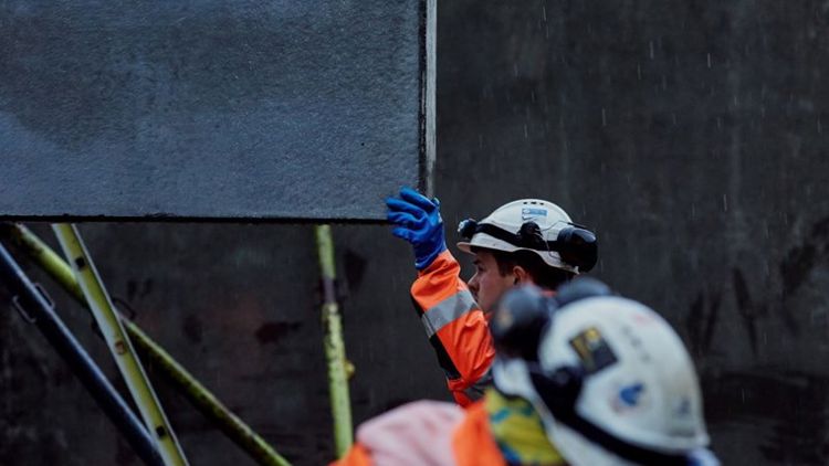 Bildet viser en byggeplass hvor to menn jobber. Vi ser at den ene mannen tar imot et element som senkes med kran.