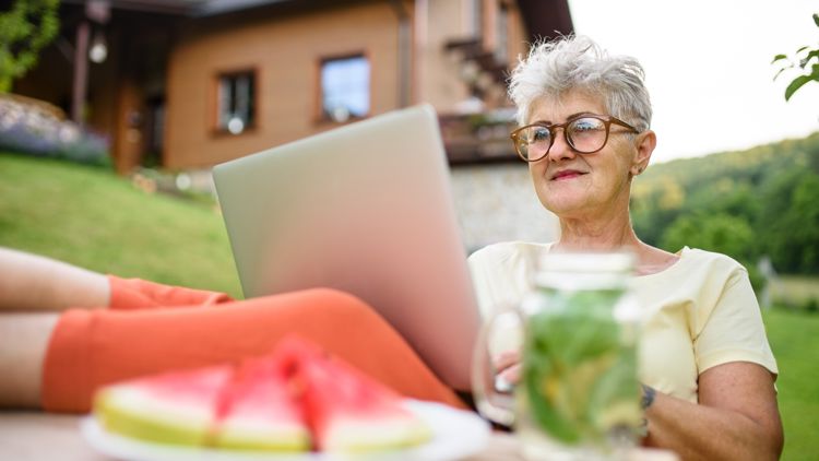 Biletet viser ei eldre dame som sit med PC på fanget ute i hagen med vassmelon på bordet.