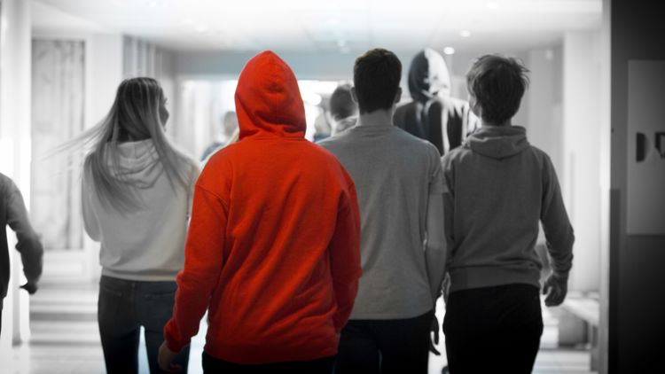 Flere skoleelever sett bakfra går bortover en gang, den ene med en rød hettegenser med hetta trukket over hodet