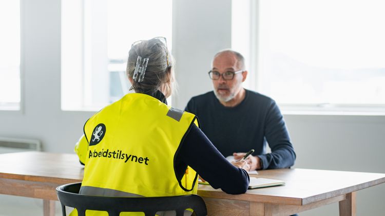en kvinnelig inspektør fra Arbeidstilsynet i gul vest med logo sitter ved et bord og snakker med en mann