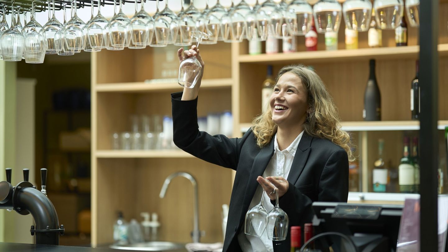 En kvinnelig bartender strekker seg opp for å hente et vinglass fra en hylle over baren. Rundt henne står flasker og annet serveringsutstyr.