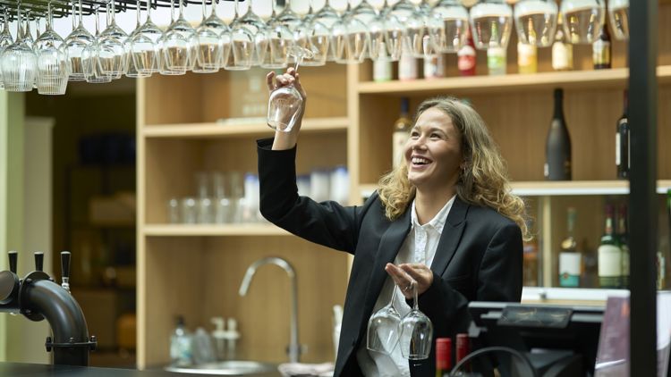 En kvinnelig bartender strekker seg opp for å hente et vinglass fra en hylle over baren. Rundt henne står flasker og annet serveringsutstyr.