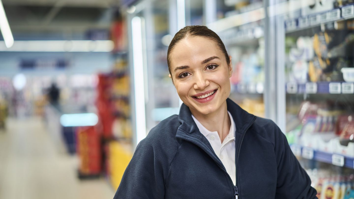  En ung, kvinnelig butikkansatt i nærbilde. Hun står inne i en butikk og smiler inn kamera.