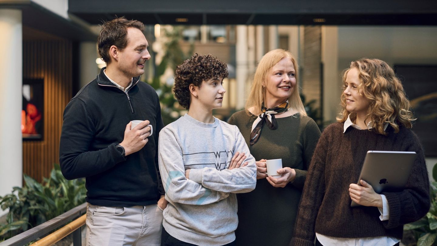  Fire mennesker står ved siden av hverandre. De tre til venstre på bildet ser på hun til høyre. Hun ser på dem. Hun holder en PC. Alle smiler og er plassert i noe som ser ut som et vrimleområde i et kontorbygg.